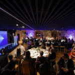 Panorámica do salón con numerosos comensais en mesas redondas e un orador no atril