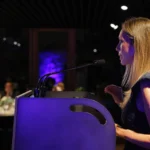 Unha muller de perfil fala ante o micrófono dun atril iluminado en violeta, co público sentado ás mesas ao fondo