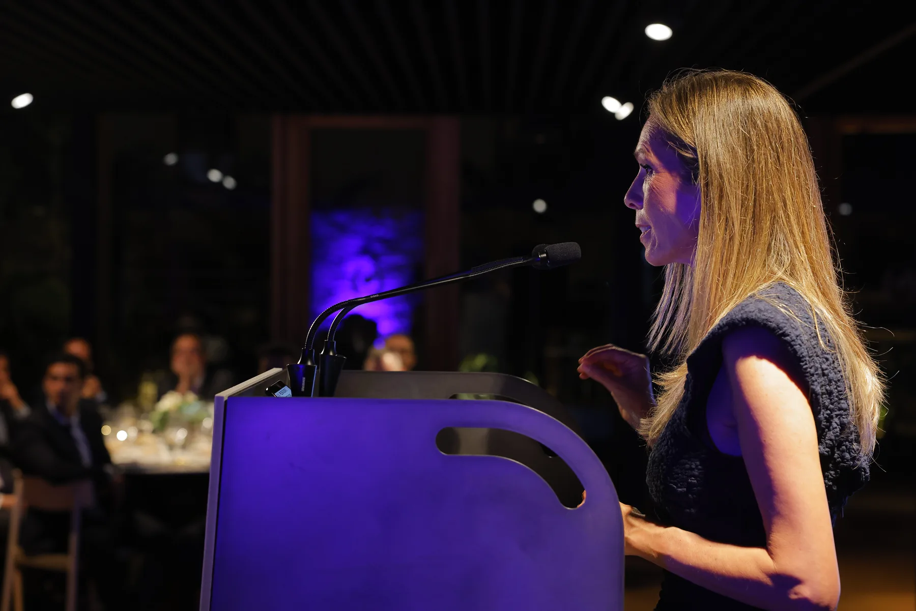 Unha muller de perfil fala ante o micrófono dun atril iluminado en violeta, co público sentado ás mesas ao fondo